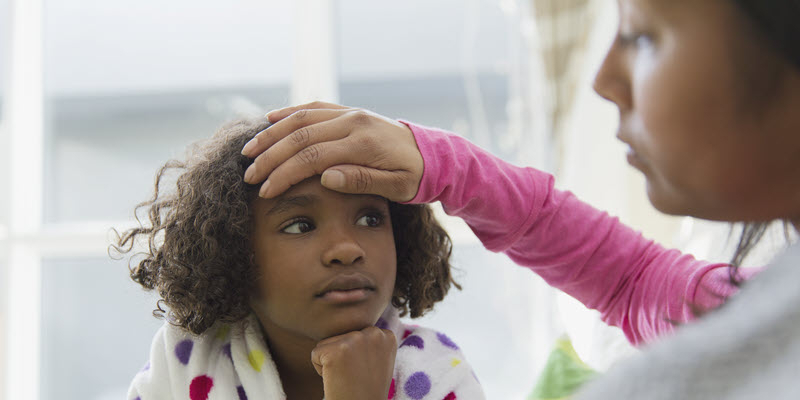 An adult checks a young child's forehead for a fever. The child is wearing a polka dot robe and looks up at the adult.