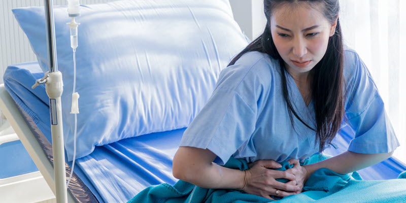Woman in a hospital gown sitting on a bed, clutching her abdomen and appearing to be in pain, with an IV stand visible beside her.