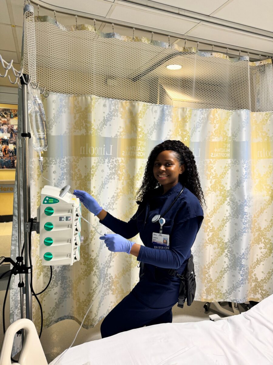 A nurse in navy scrubs and gloves stands beside a hospital bed, adjusting an IV machine in a patient room with privacy curtains.