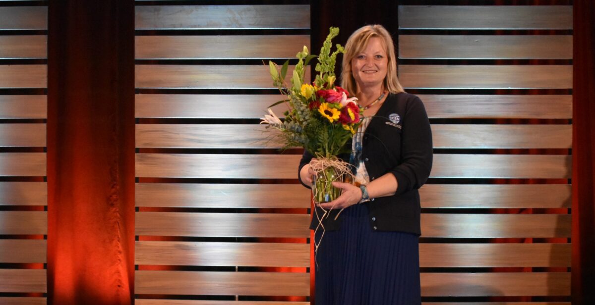 A woman stands on stage in front of wooden panels, holding a large bouquet of flowers and smiling at the camera.