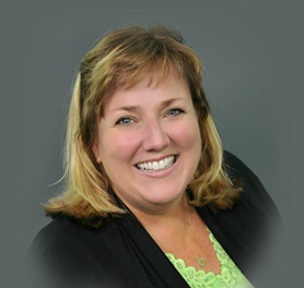 A woman with blonde hair wearing a black jacket and light green top smiles in front of a gray background.