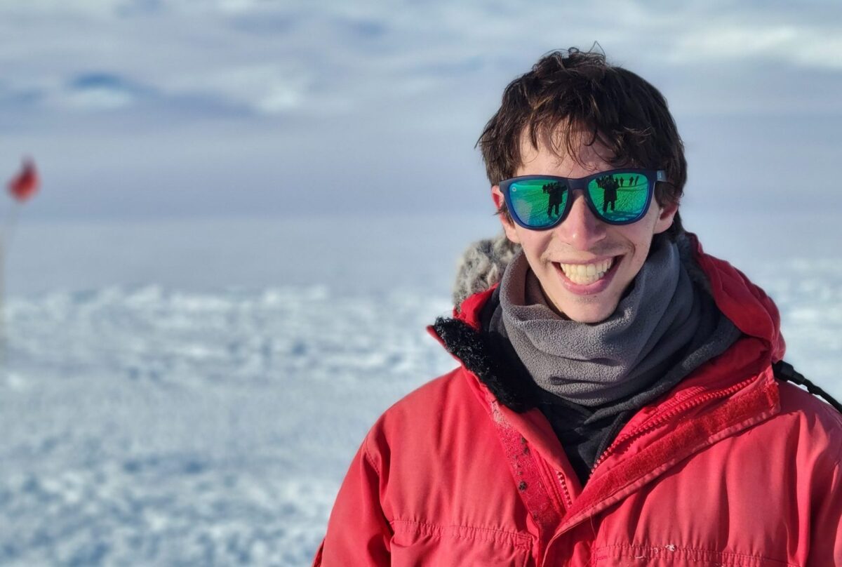 Person wearing a red winter jacket, gray scarf, and reflective sunglasses smiling outdoors in a snowy, icy landscape.