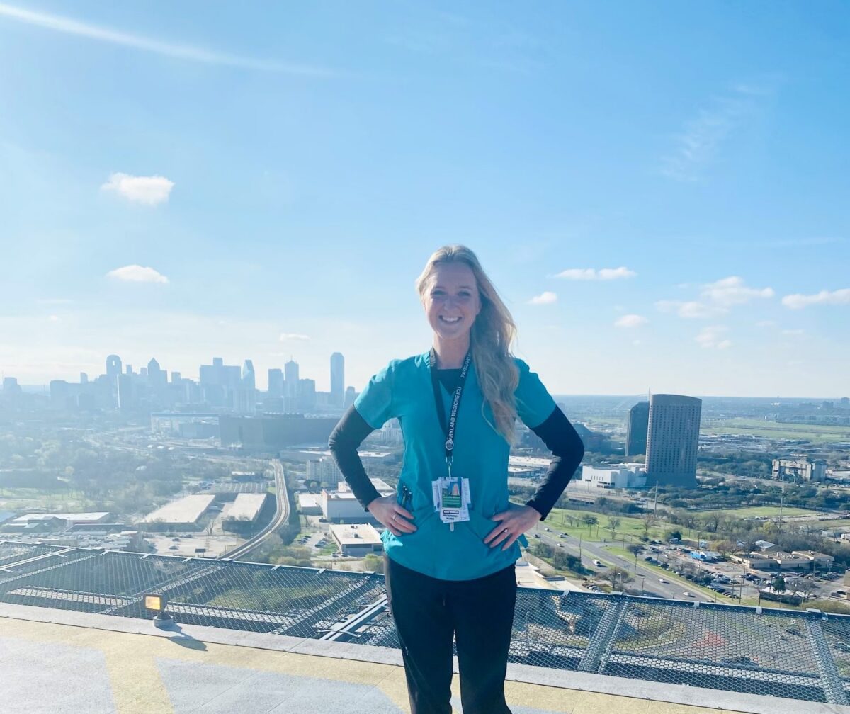 A woman in medical scrubs stands on a rooftop helipad with a city skyline and blue sky in the background.