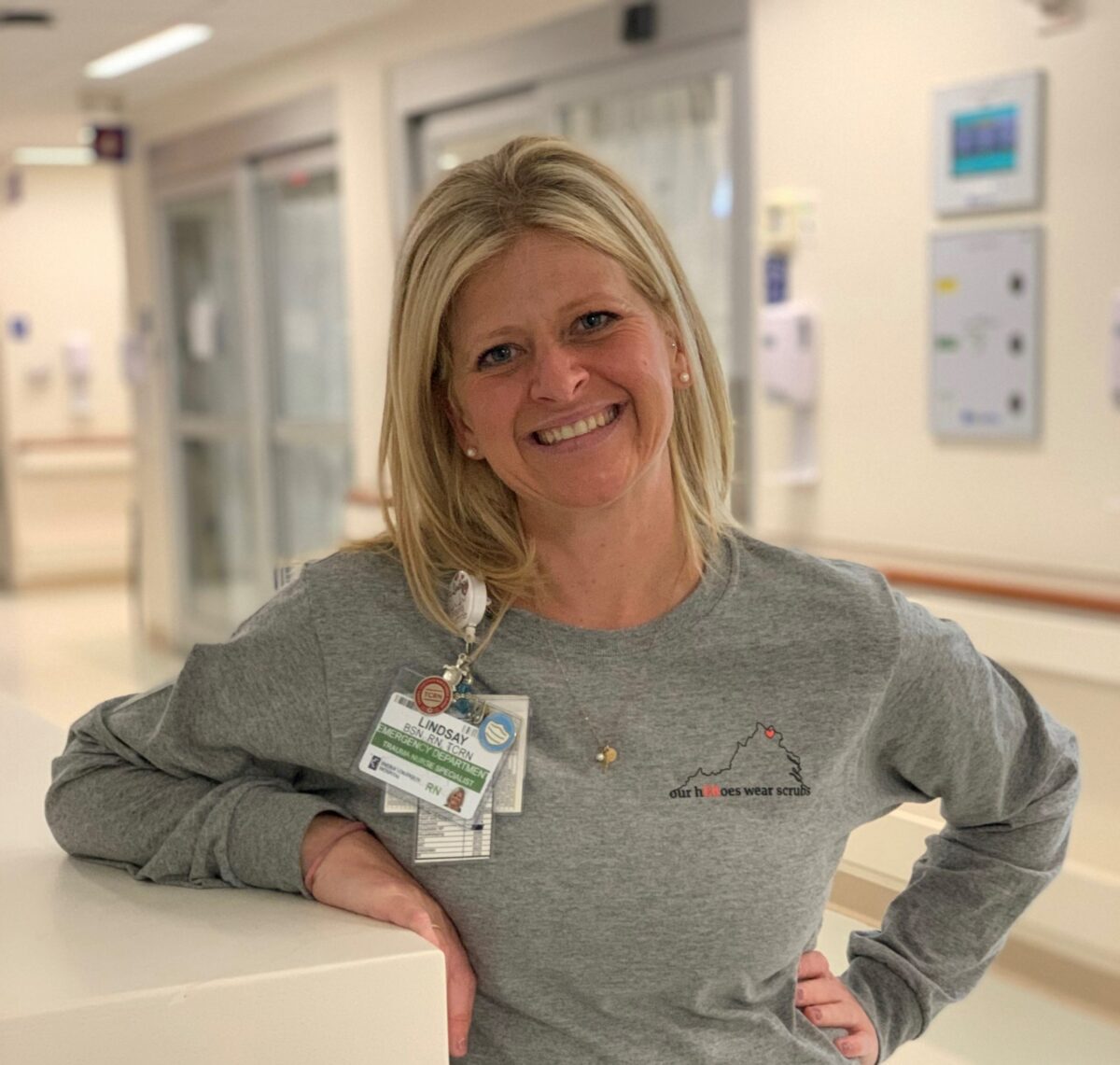 A woman with blonde hair wearing a gray shirt and hospital badges smiles and leans on a counter in a hospital corridor.