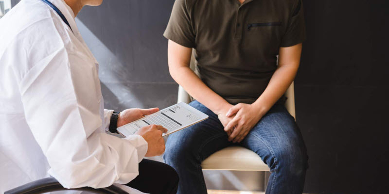 A doctor holds a clipboard and talks with a seated man in casual clothes during a consultation.