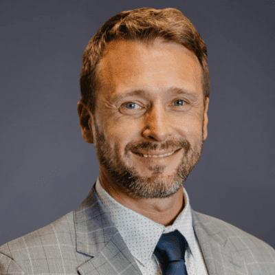A man with short light brown hair and a beard, wearing a light gray checked suit, white shirt, and dark blue tie, smiling in front of a plain dark background.