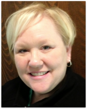 A woman with short blonde hair, smiling, wearing small hoop earrings and a black top, posed in front of a wooden background.