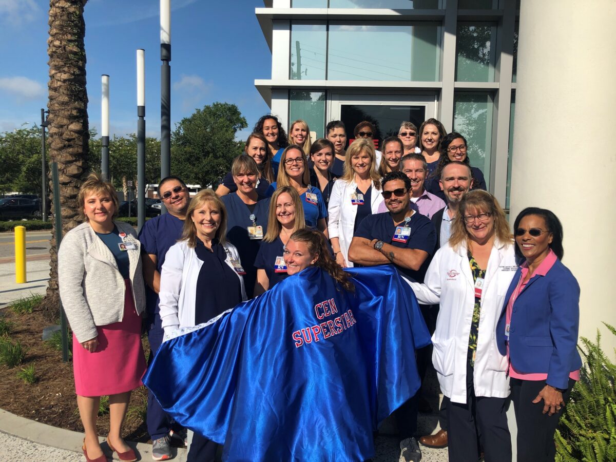 A group of healthcare professionals poses outside a building; one person stands in front wearing a blue cape labeled "CEN Superstar.