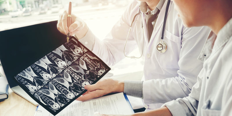 Two doctors review and discuss medical scan images on a film while sitting at a desk with paperwork and a laptop.