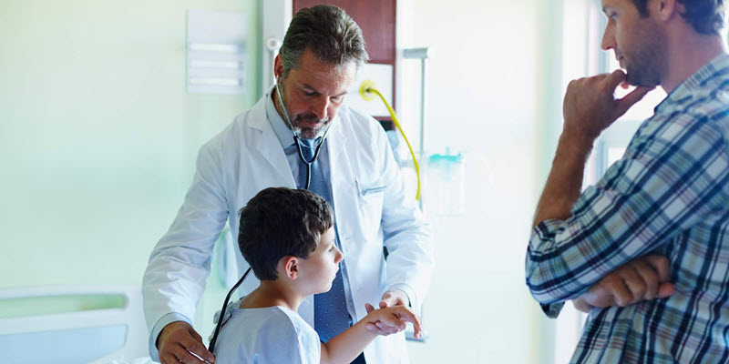 A doctor examines a young boy with a stethoscope in a medical room while an adult stands nearby observing.