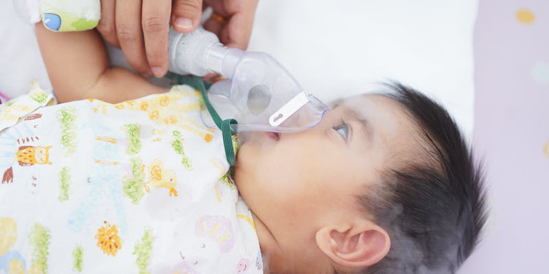 A baby lying down wears a nebulizer mask while an adult hand holds the device, with visible mist coming from the mask.