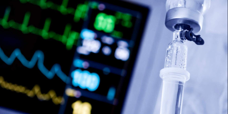 Close-up of an IV drip with a blurred hospital monitor displaying vital signs in the background.