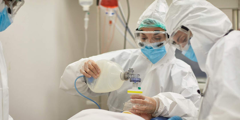 Three healthcare workers in full protective gear and masks are administering oxygen to a patient using a manual resuscitator in a medical setting.