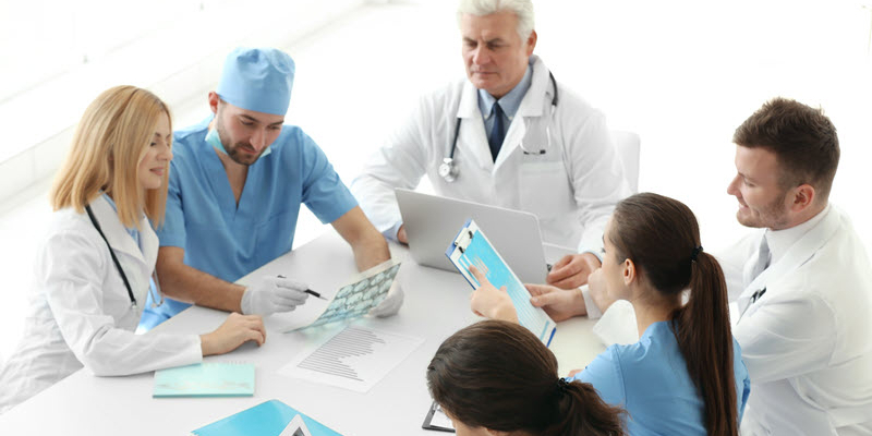 A group of medical professionals sit around a table reviewing documents and X-rays during a meeting.