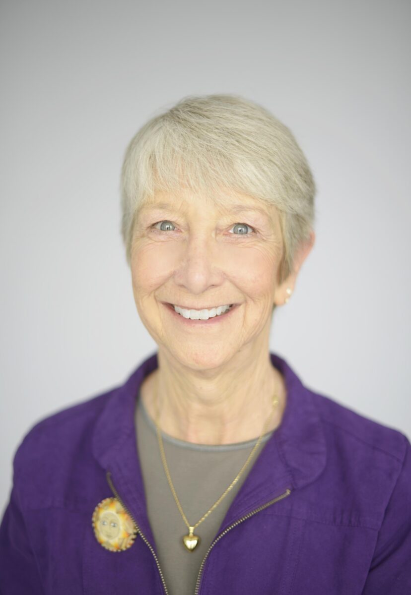 Older woman with short gray hair, smiling, wearing a purple jacket, gray top, and gold jewelry, standing against a plain light background.
