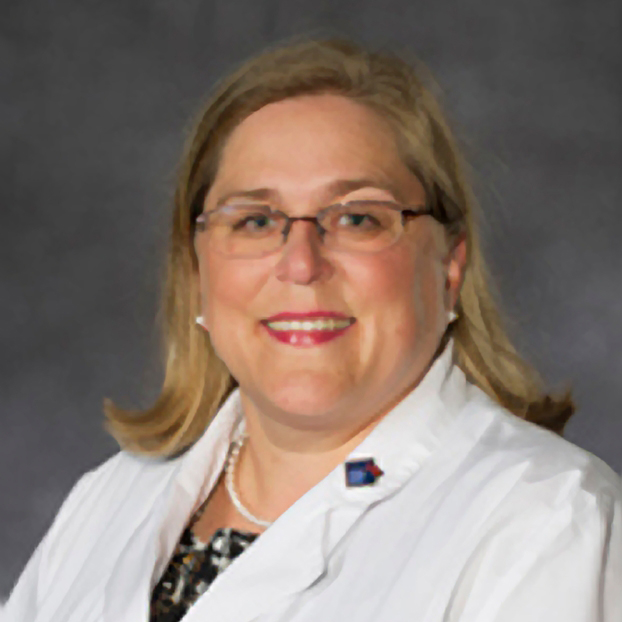 A woman with long blonde hair, wearing glasses, a white lab coat, and a pin, poses in front of a plain gray background.