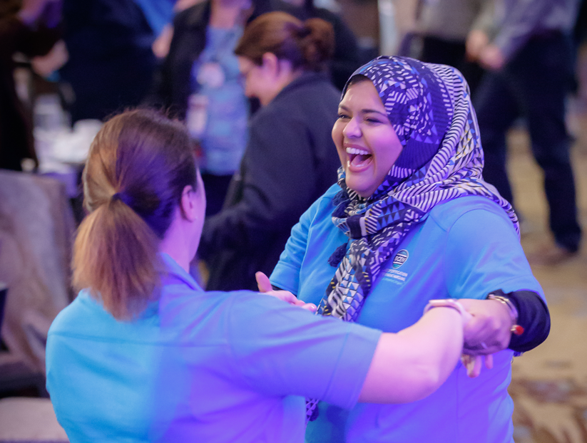 Two women in blue shirts, one wearing a patterned hijab, smile and hold hands while interacting at a busy indoor event.