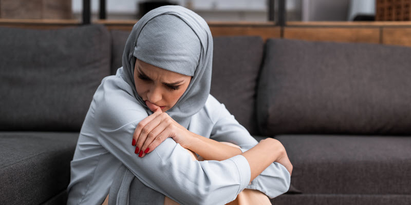 A woman wearing a grey headscarf sits on a couch with her arms folded over her knees, looking down with a concerned expression.