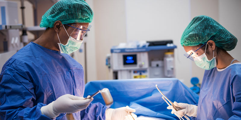 Two surgeons in blue scrubs, masks, and green caps perform a surgical procedure in an operating room with medical equipment in the background.