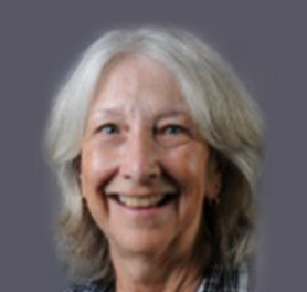 An older woman with gray hair and a friendly smile, wearing a plaid shirt, is pictured against a plain gray background.