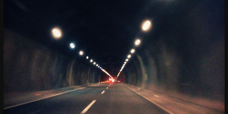 View from inside a tunnel with illuminated lights along the ceiling and a car visible in the distance on the road ahead.
