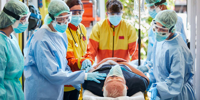Medical team in protective gear attends to an elderly patient lying on a stretcher, possibly in an emergency or hospital setting.