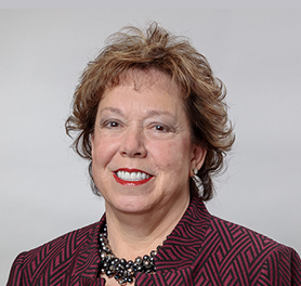 Woman with short, curly brown hair, wearing a patterned maroon jacket, a beaded necklace, and smiling in front of a plain light gray background.