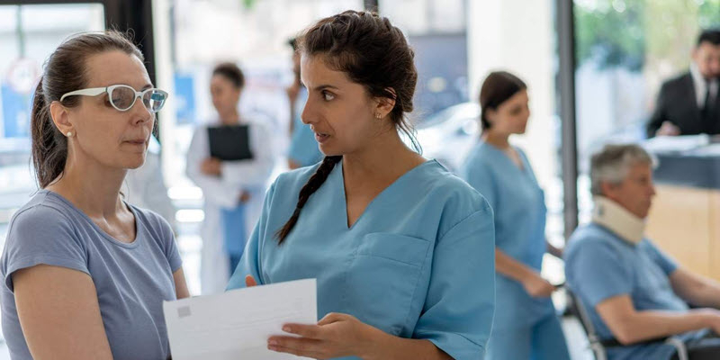 A nurse speaks with a woman holding paperwork in a busy medical waiting area with other staff and patients visible in the background.