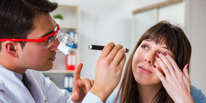 A doctor examines a woman's eye using a flashlight while she holds her hand near her eye in a medical office setting.
