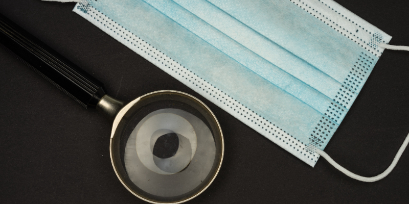 A blue surgical face mask and a magnifying glass are placed on a black surface.