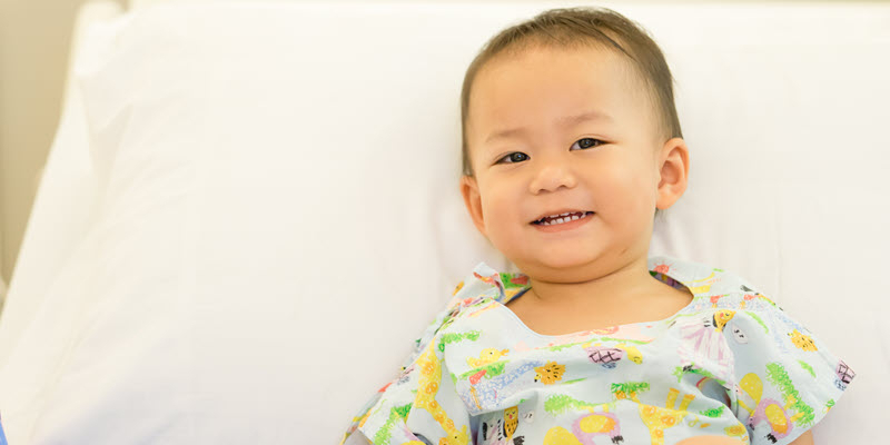 A young child wearing a patterned hospital gown smiles while sitting up in a hospital bed.
