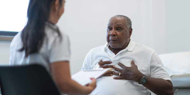 An older man sits on a medical exam table, holding his chest while speaking to a healthcare professional taking notes.