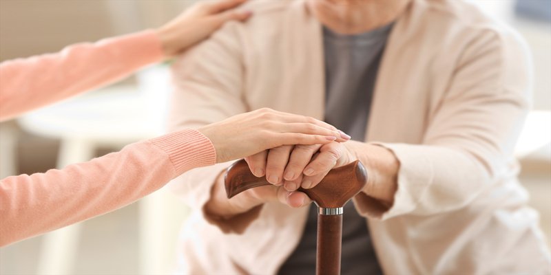 Close-up of a younger person’s hand comforting an older person who is holding a cane. Both individuals are wearing light-colored clothing.