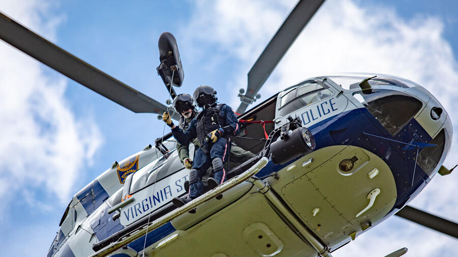 Two people in tactical gear prepare to rappel from a Virginia State Police helicopter in flight against a partly cloudy sky.