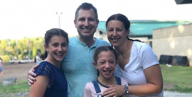 Four people, two adults and two children, stand close together outdoors, smiling at the camera on a sunny day.