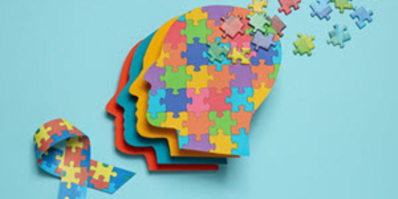A layered silhouette of human heads made from colorful jigsaw puzzle pieces, with some pieces floating away at the top, next to a ribbon of puzzle pieces.