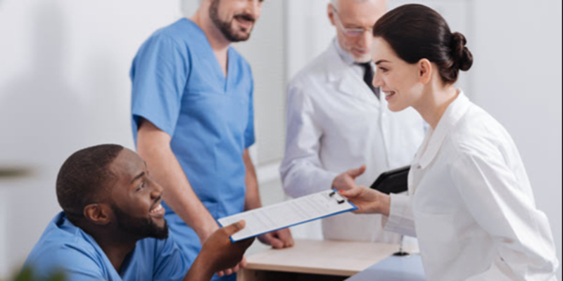 Three healthcare professionals stand in discussion while a fourth person, seated and wearing scrubs, hands a clipboard to a standing colleague in a white coat.