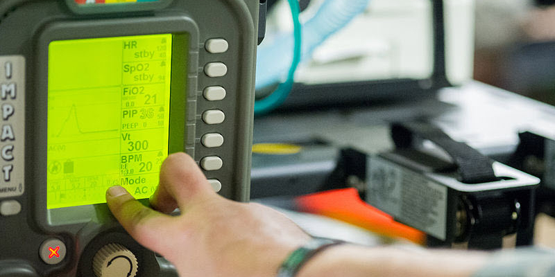 A hand adjusts settings on a medical monitor displaying heart rate, oxygen saturation, and breathing parameters.