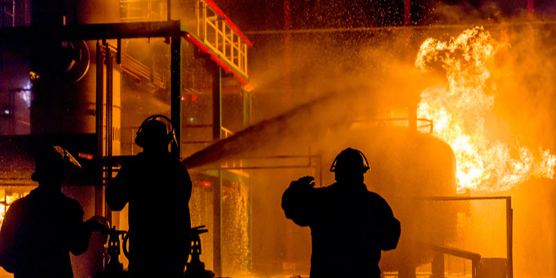 Three firefighters in silhouette spray water at a large fire engulfing an industrial structure at night.