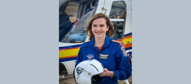 A woman in a blue flight suit stands in front of a helicopter, holding a white helmet and smiling at the camera.