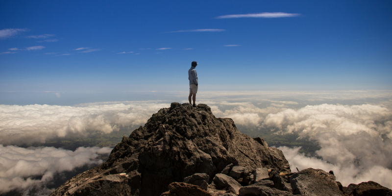 A person stands on a rocky mountain peak under a blue sky, looking out over a sea of clouds below.
