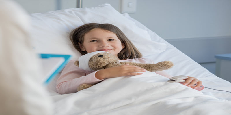 Young girl lying in a hospital bed, smiling and holding a teddy bear, with a pulse oximeter on her finger.