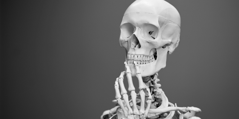 A human skeleton model with one hand touching its chin, posed as if thinking, against a plain gray background.