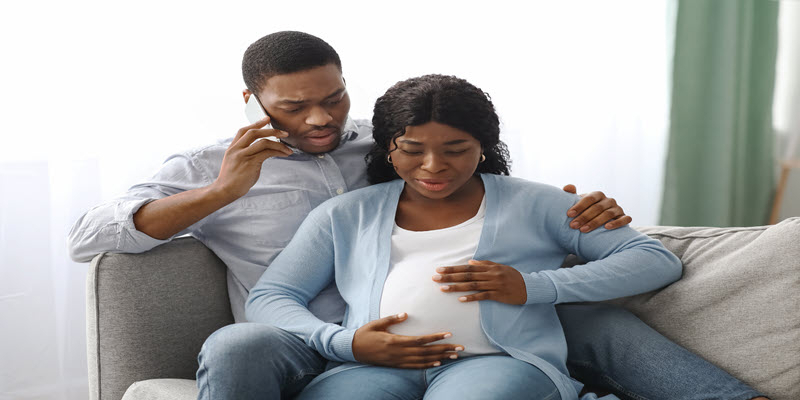 A man talks on the phone while sitting beside a pregnant woman on a couch, both looking concerned with the woman's hand on her belly.