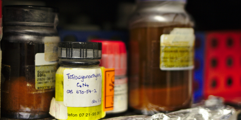 Several glass and plastic chemical containers, some with handwritten and faded labels, are arranged on a shelf in a laboratory setting.