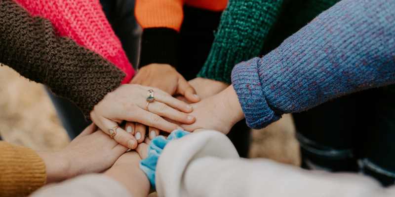 A group of people wearing colorful sweaters place their hands together in the center, showing teamwork and unity.