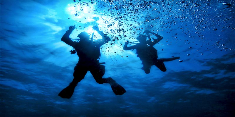 Two scuba divers swim underwater surrounded by bubbles, with sunlight filtering through the surface above them.