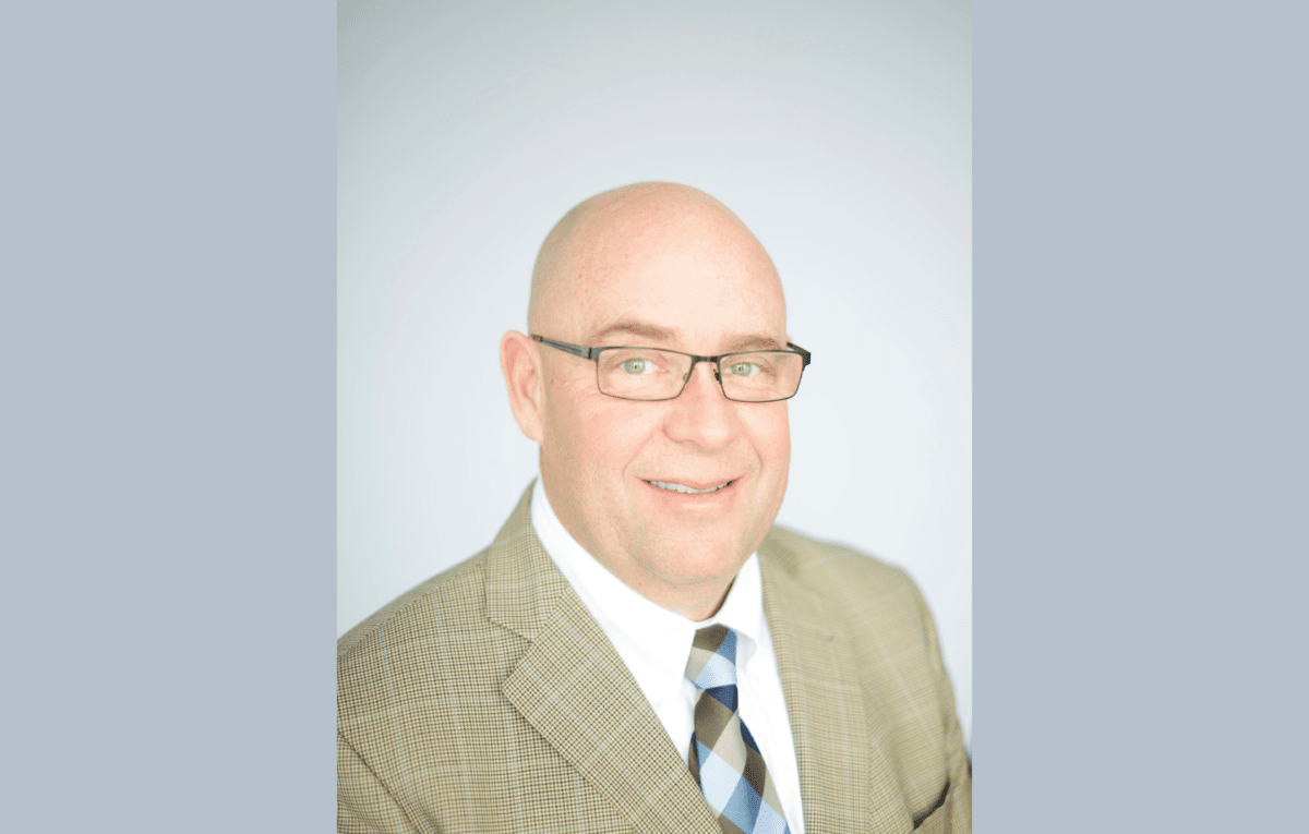 A bald man wearing glasses, a tan plaid suit jacket, white shirt, and striped tie, smiling in front of a plain light background.