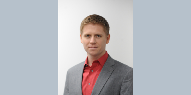 A man with short light brown hair wearing a grey suit jacket and a red collared shirt stands in front of a plain white background.