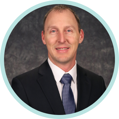 A man in a dark suit, light blue shirt, and blue tie poses for a professional headshot against a textured dark background.